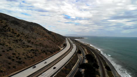 aerial drone shot over the 101 freeway with cars next to the blue pacific ocean waves and california coast cliffs in ventura