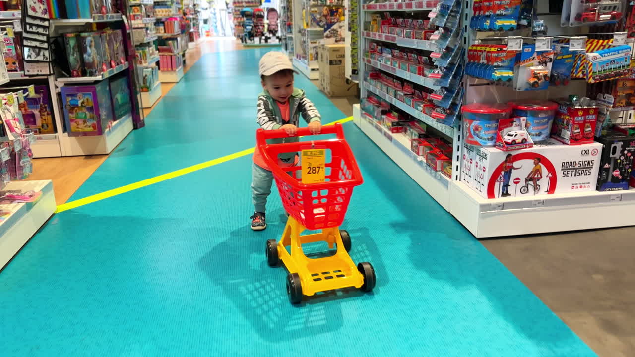 Excited baby boy pushing the red cart by the store isle. Cute child shopping in a toy shop.