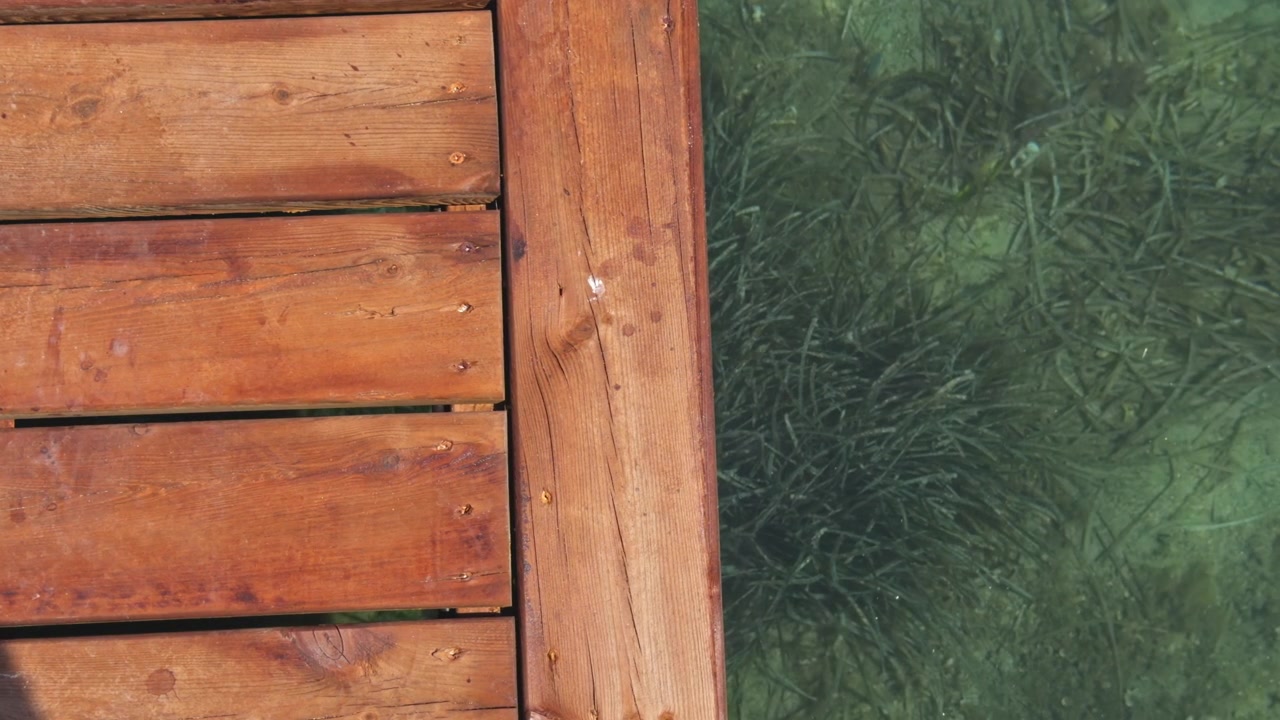 Top-down overhead shot of wooden pier edge over crystal clear Aegean water, seagrass visible on seabed below, beach club pier detail, Turkey