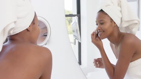 Happy-biracial-woman-with-towel-on-head-looking-in-mirror-washing-her-face,-slow-motion
