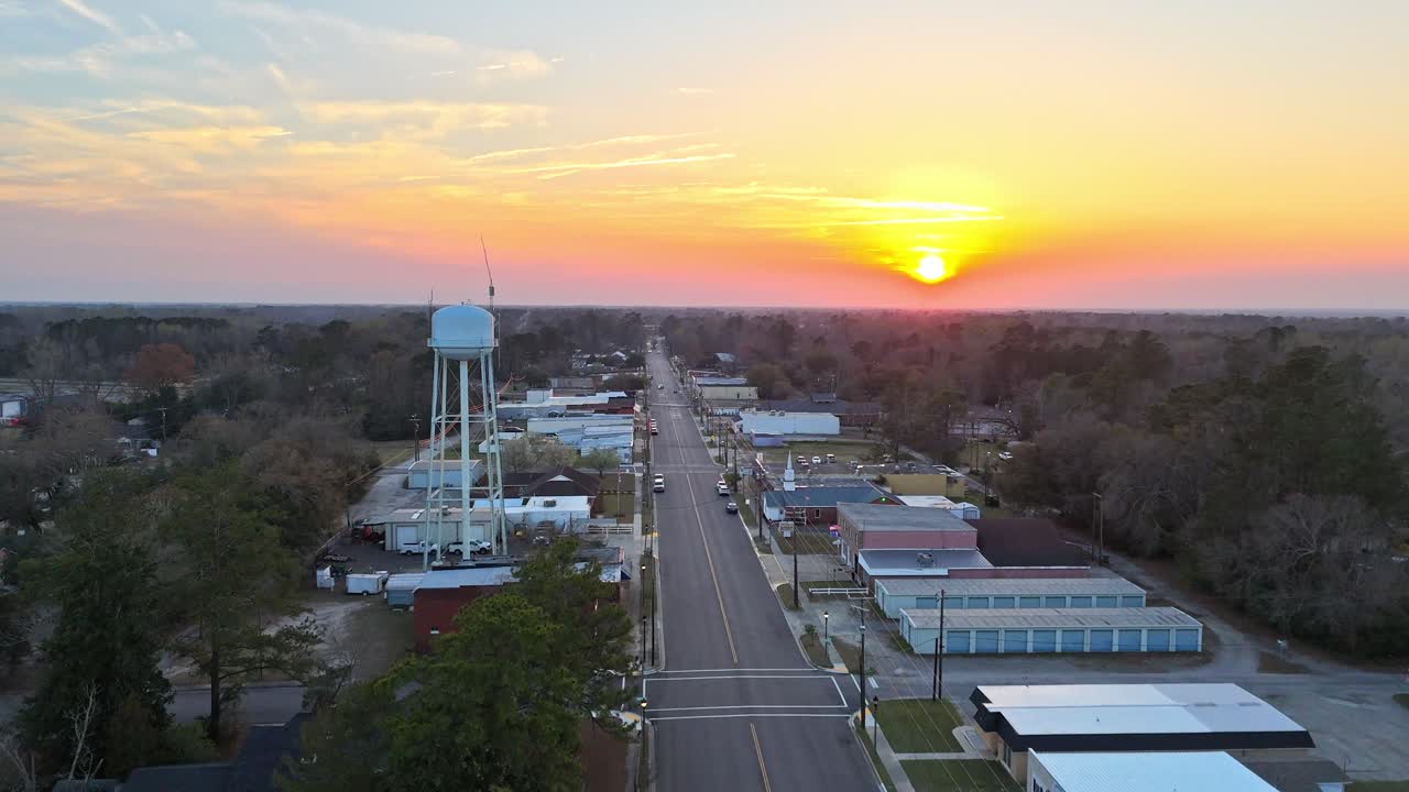 A reversing drone shot of the main street in downtown Johnsonville, SC showing cars, buildings, and trees. A bright sunset fills the sky with orange and yellow color in the background.