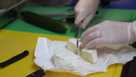 la mano del chef con el guante cortando queso brie redondo usando un cuchillo en la tabla de cortar