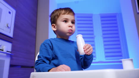sweet baby boy wearing blue sweater sits at high chair drinking yogurt from the bottle. low angle view at the kid enjoying dairy at home.