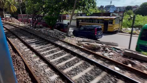 pov shot from a moving train in colombo, sri lanka, showcasing the cityscape and railway tracks alongside a road with buses and buildings