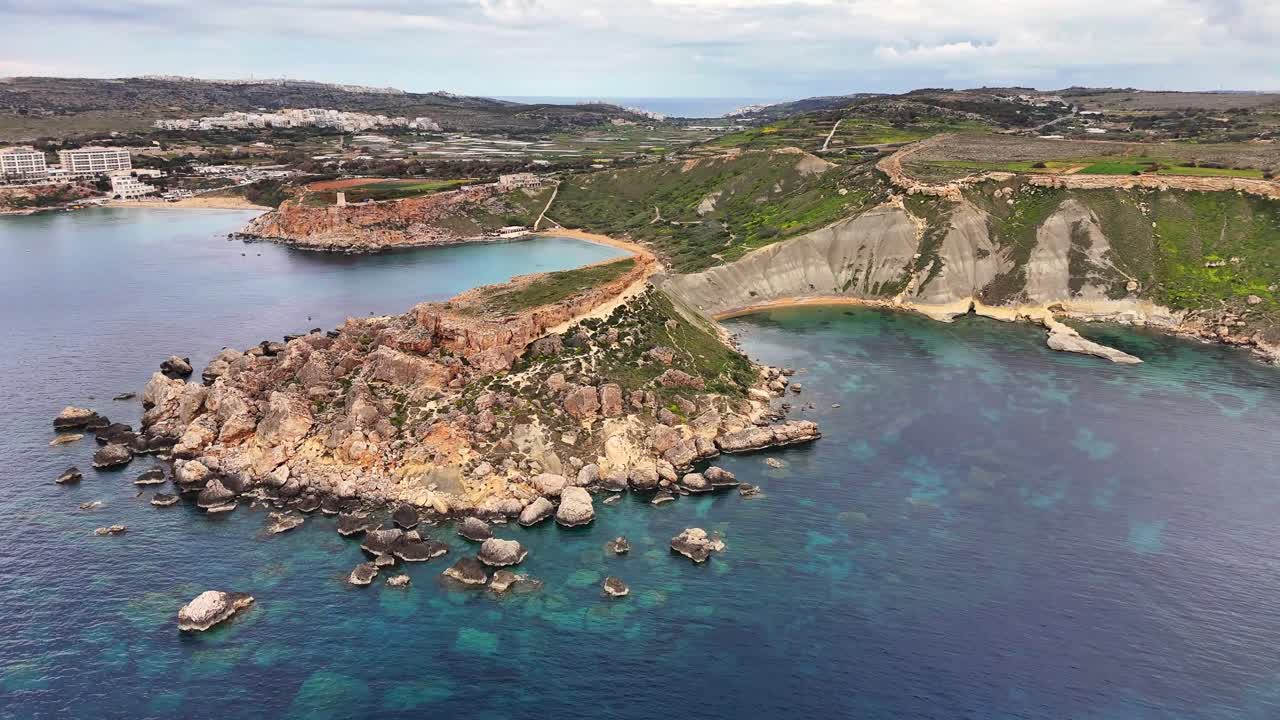 Baia di Qarraba a Malta che mostra un promontorio calcareo e pendii di argilla blu modellati da processi di frana sopra acque limpide del Mediterraneo all'interno del sistema della Baia di Ġnejna, ripresa fluida con drone in allontanamento