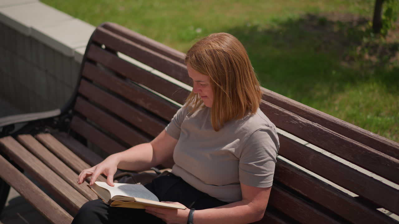 Ragazza seduta su una panchina di legno che legge un libro all'aperto in una giornata di sole, rilassata e concentrata mentre sfoglia le pagine, con auto che passano lentamente sullo sfondo sfocato e foglie verdi che ondeggiano