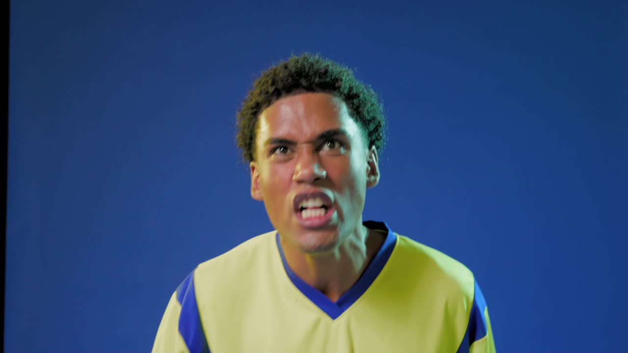 Adult African male in yellow jersey shouting after cue thrusting arm in blue studio holding whistle