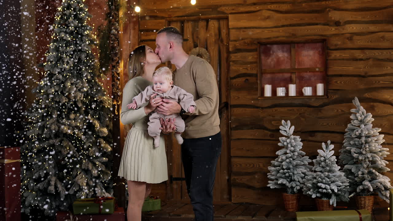 Loving couple with their beloved baby boy. Happy parents of an infant kissing under the falling snow. Christmas backdrop.