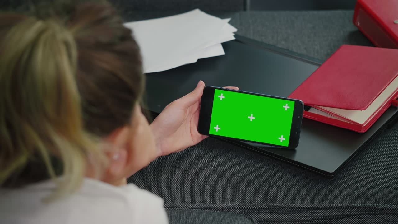 mujer sentada en un sofá con teléfono inteligente en modo horizontal con pantalla verde simulada de pantalla de clave de croma.