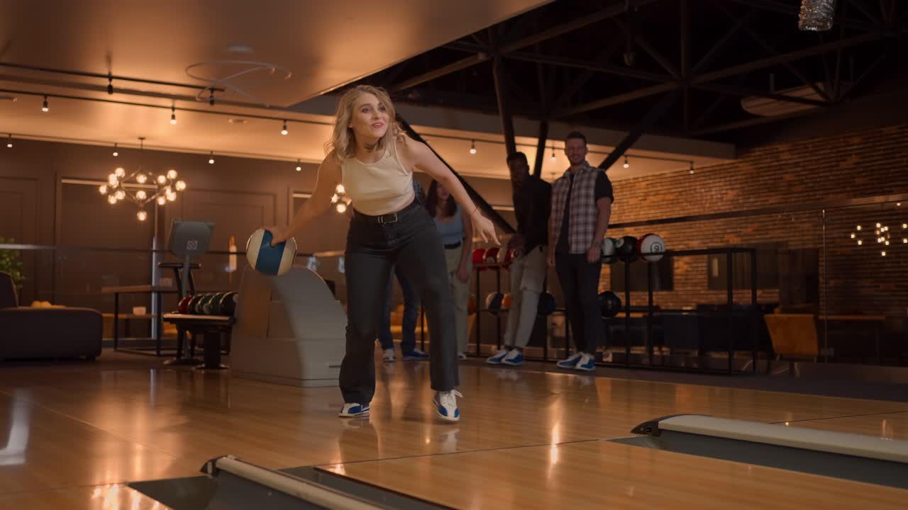Una giovane donna al bowling lancia una palla sulla pista e cade a terra al rallentatore, poi salta e balla di gioia. Gli amici fan la supportano e applaudono. Amici multietnici giocano a bowling insieme.