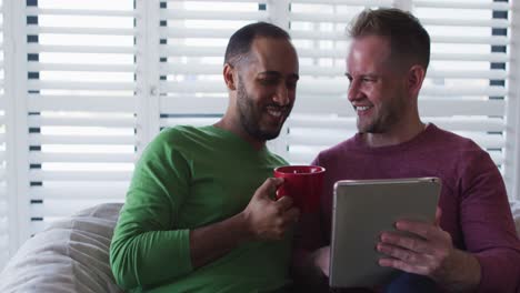 Multi-ethnic-gay-male-couple-sitting-on-couch-one-using-tablet