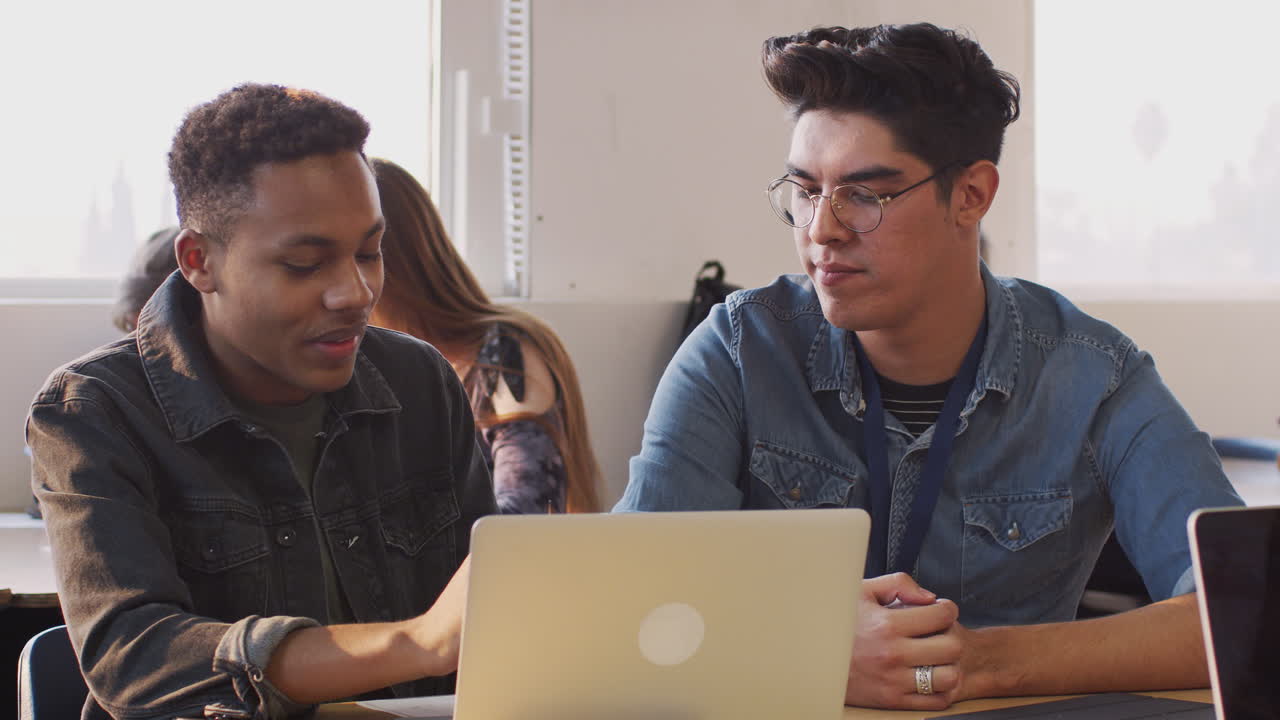 Male Teacher Giving One To One Support To Student Working At Desk On Laptop