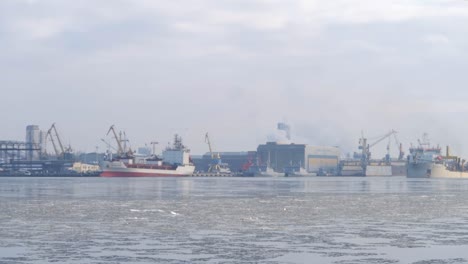 industrial port with cargo ships and cranes in the distance with frozen ice cold water in between on an overcast day