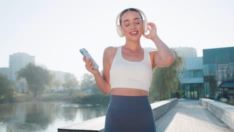 young runner girl holding smartphone listening music on headphones dancing having fun in city park