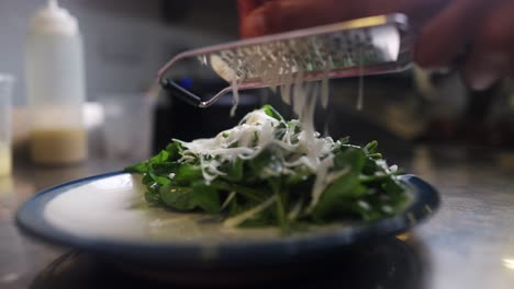 close up view of a chef sprinkling grated cheese over a mix of lettuce in a restaurant cuisne