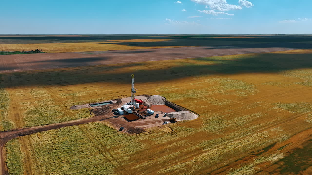 Flying away from the gas producing site located in the vast field. Shadows from clouds are on the ground.