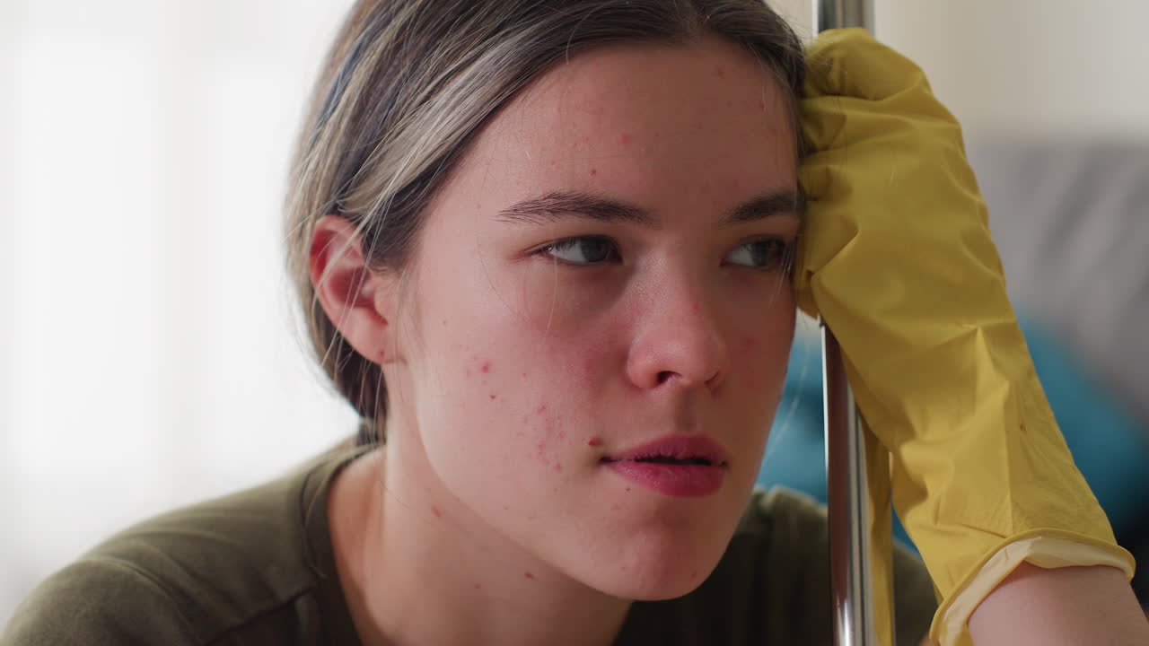 Close up of girl in yellow gloves holding mop handle with eyes closed looking frustrated exhausted from cleaning chores showing fatigue stress burnout tired expression during household work routine