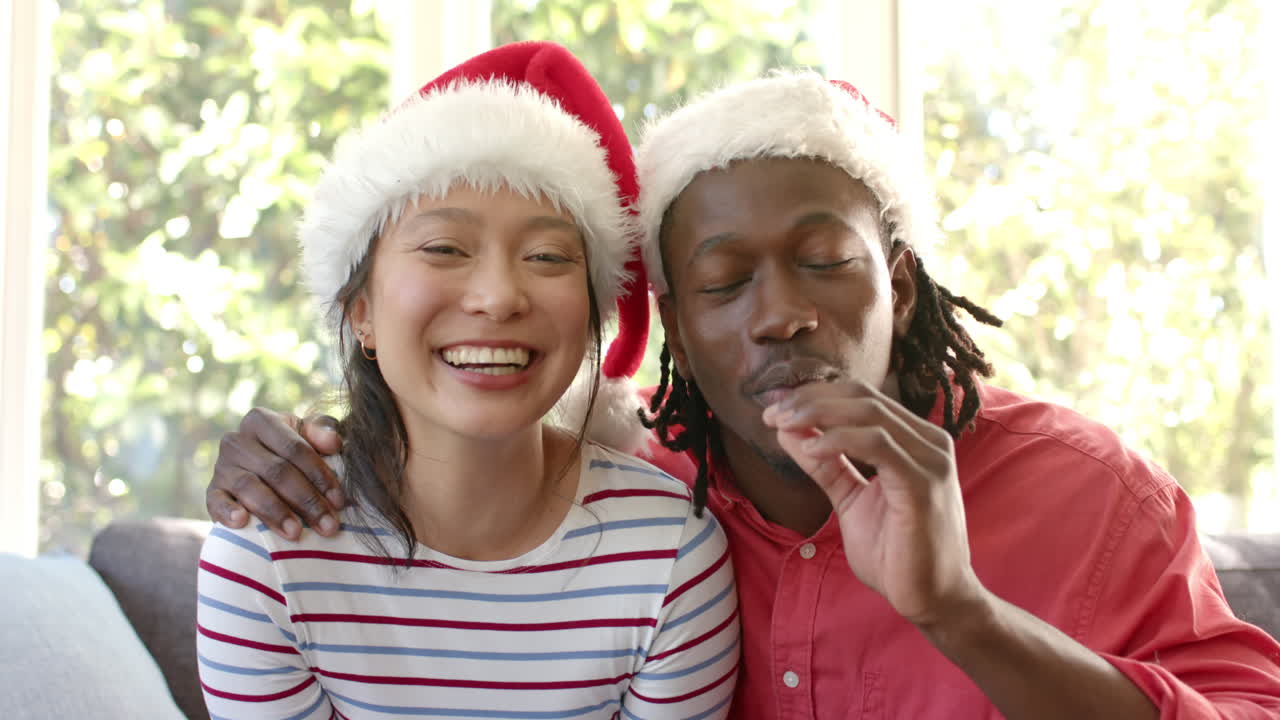 una pareja feliz y diversa con sombreros de navidad haciendo una llamada de video en una sala de estar soleada, en cámara lenta.