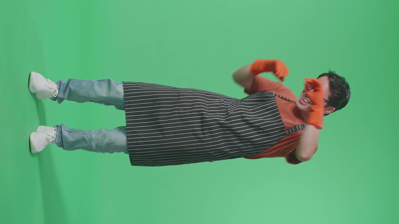 Full Body Of Cheerful Asian Male Housekeeper With An Apron Dancing While Standing In The Green Screen Studio