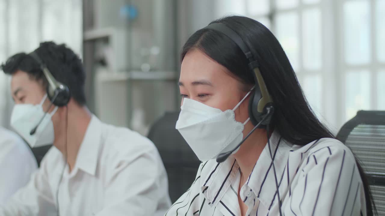 Close Up Of An Asian Woman Call Center Agent Wearing Headset And Mask Speaking To Customer On The Call During Working With Colleagues At The Office