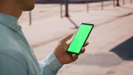 close up of a young man using smartphone with green screen chroma key mock up display in vertical position on a street during a sunny day. he's tapping and swiping the screen.