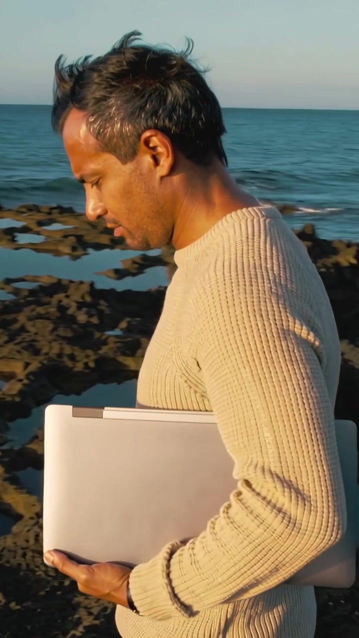 Video verticale: Uomo che cammina su una costa rocciosa tenendo un laptop per lavoro, indossando un capo in maglia beige.
