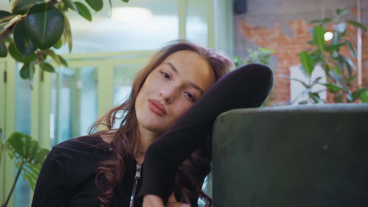 Confident woman with soft expression leans on cushion and gazes at camera in warmly decorated indoor space with green plants and blurred background, evoking calm and poised ambiance