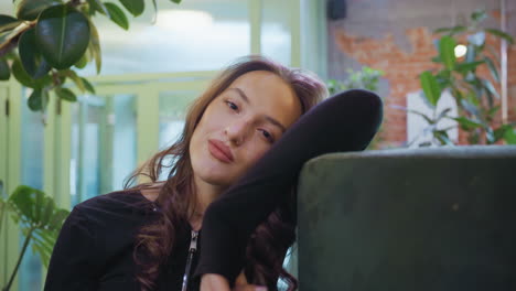 confident woman with soft expression leans on cushion and gazes at camera in warmly decorated indoor space with green plants and blurred background, evoking calm and poised ambiance