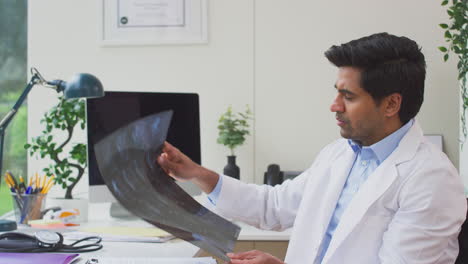 Male-Doctor-Wearing-White-Coat-Sitting-At-Desk-Looking-At-CT-Or-MRI-Scan