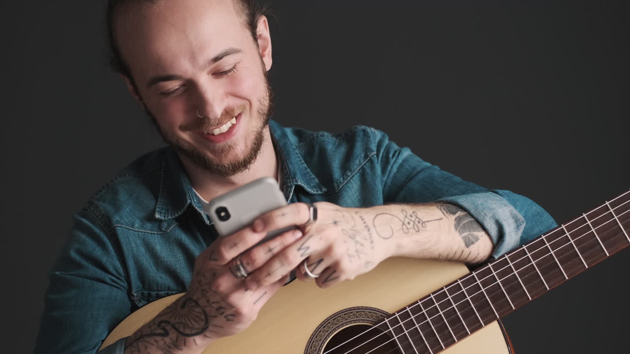 un joven caucásico enviando mensajes de texto en su teléfono inteligente mientras sostiene una guitarra.