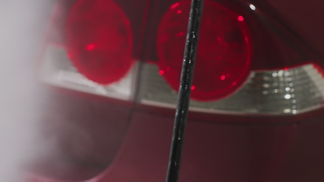close up of red car tail lamp being rinsed by high pressure water spray inside garage with blurred mist foam capturing glistening droplets on glossy metal and plastic surfaces a
