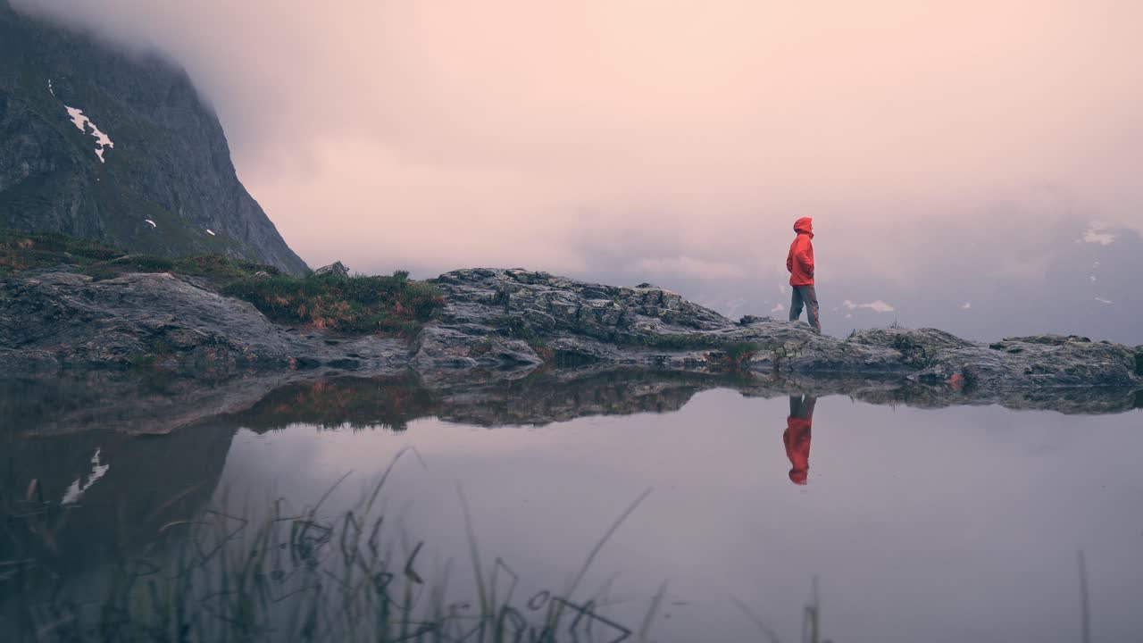 Osoba idąca po skalistej górze, obok swojego odbicia w wodzie, otoczona górami i mglistymi chmurami. Norwegia, Litlefjellet.