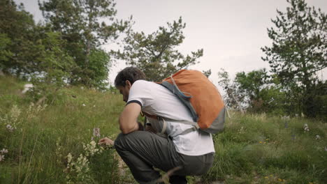 hiker with an orange backpack walking up a green path where among grass grow some meadow flowers and are blooming