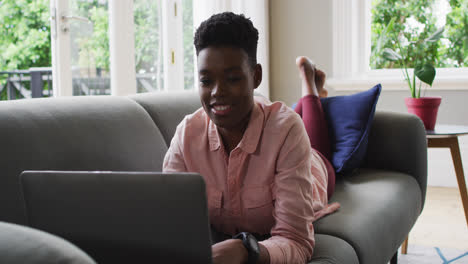mujer afroamericana usando una computadora portátil mientras yacía en el sofá en casa