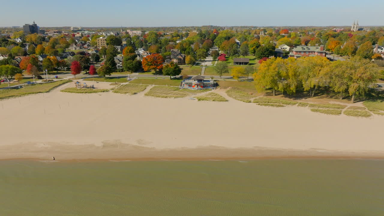 Drone aerial trucking right along the Lake Michigan shoreline in Sheboygan, Wisconsin, with neighborhood houses, colorful autumn trees, and a clear blue sky