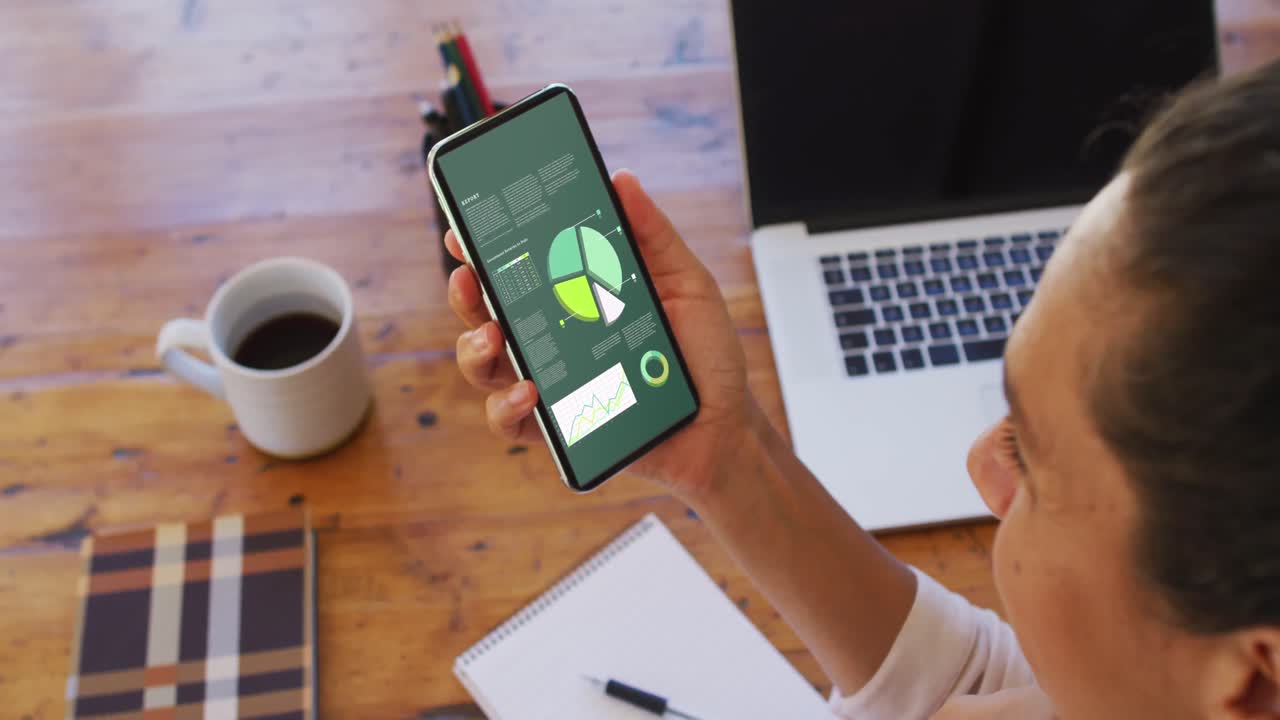 mujer de raza mixta sentada en un escritorio usando un teléfono inteligente con estadísticas en la pantalla