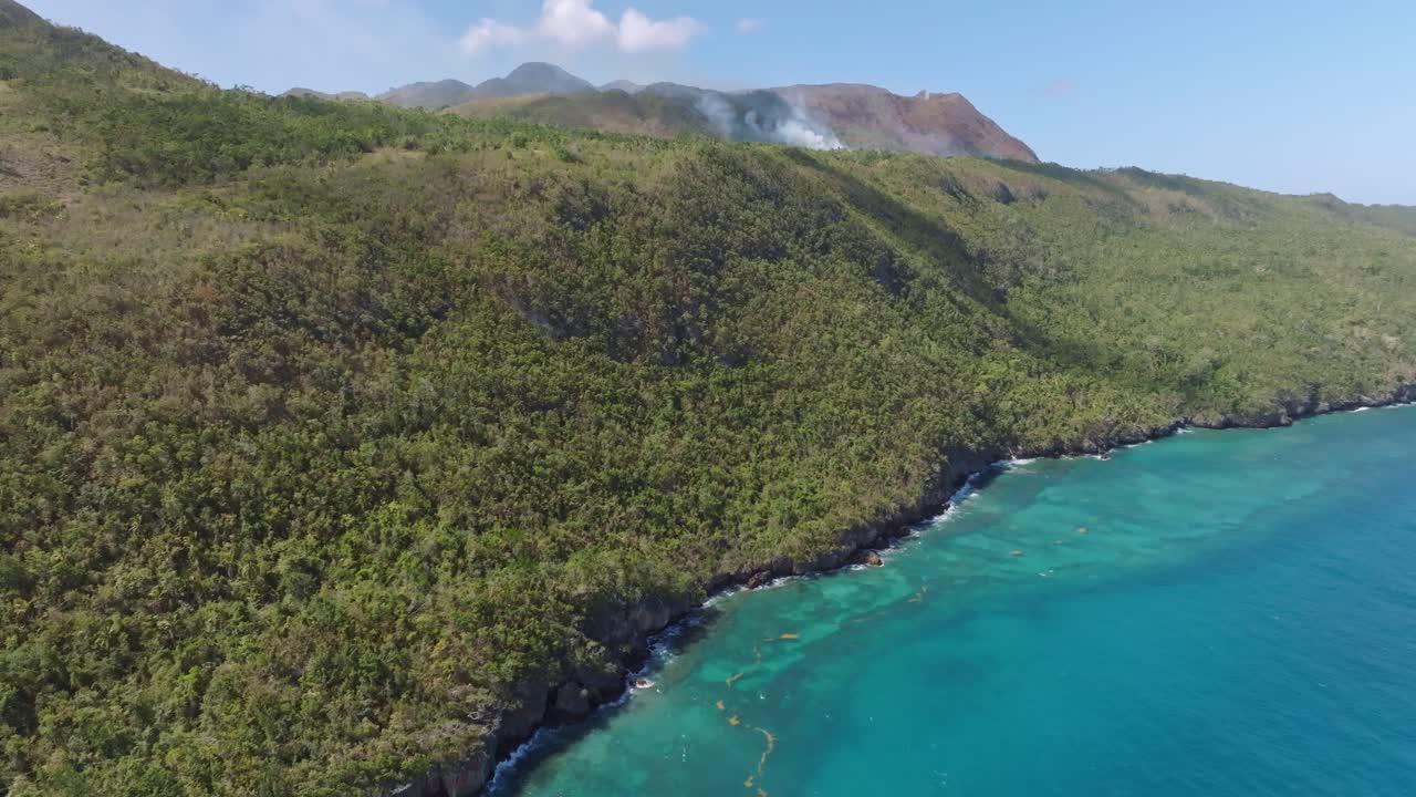 vuelo aéreo que muestra agua turquesa del mar del caribe, colinas verdes y humo de incendios forestales en el fondo - parque nacional cabo cabron, samaná