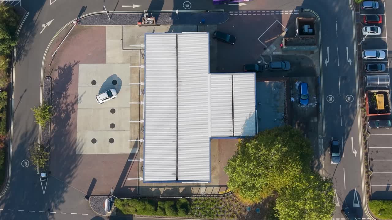 Aerial timelapse of gas station and car wash in morning light. Cars enter and exit while a person washes their vehicle. Long September shadows stretch from trees and buildings, adding depth and drama