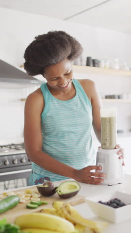 vertical video of african american woman smiling and preparing healthy drink in kitchen
