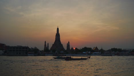 Wide-shot-of-Wat-Arun-at-sunset,-and-boats-crossing-in-front-chao-phraya-river