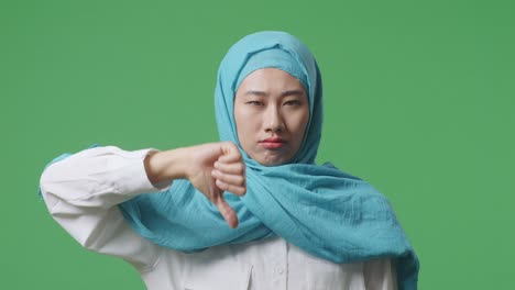 녹색 스크린 배경 스튜디오에 서 있는 아시아 무슬림 여성이 엄지손가락을 아래로 내려다보며 카메라에 제스처를 하고 있다.