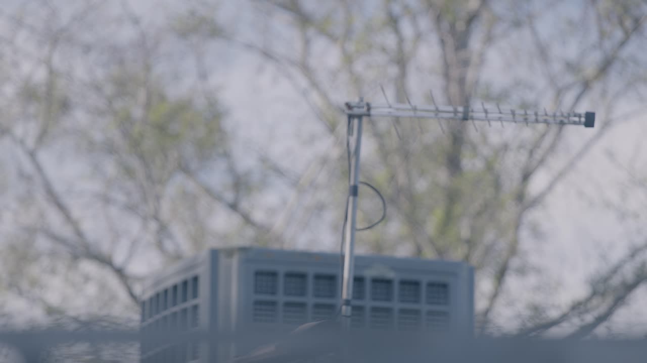An old-fashioned television antenna sits on a rooftop against a backdrop of trees and an overcast sky. This nostalgic, ungraded LOG footage is perfect for a retro aesthetic