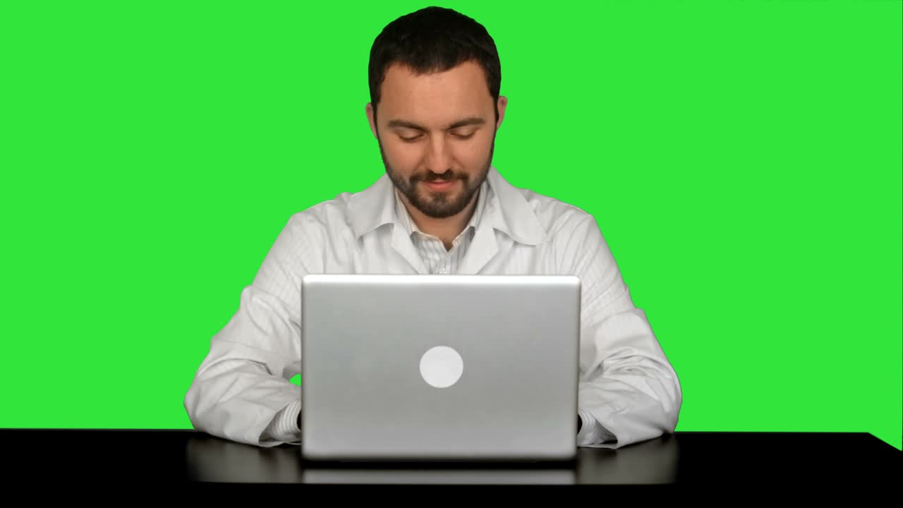 un médico de sexo masculino sonriente sentado en la mesa con una computadora portátil en una pantalla verde, llave de croma