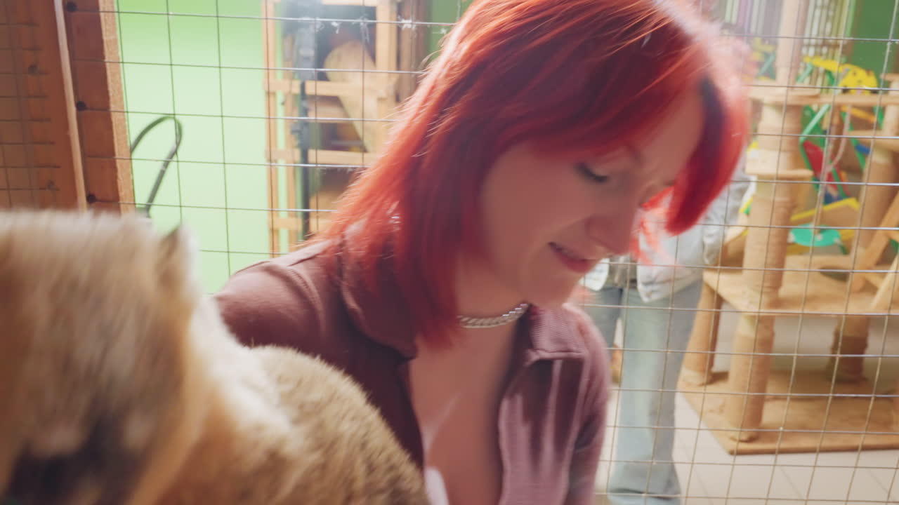 Visitante caucasiano segurando um quati perto da tela, cabelo ruivo e colar, sorrindo para o animal, vestindo roupas casuais, mãos apoiando a barriga e as patas, ambiente animado de exposição, pessoas observando ao fundo