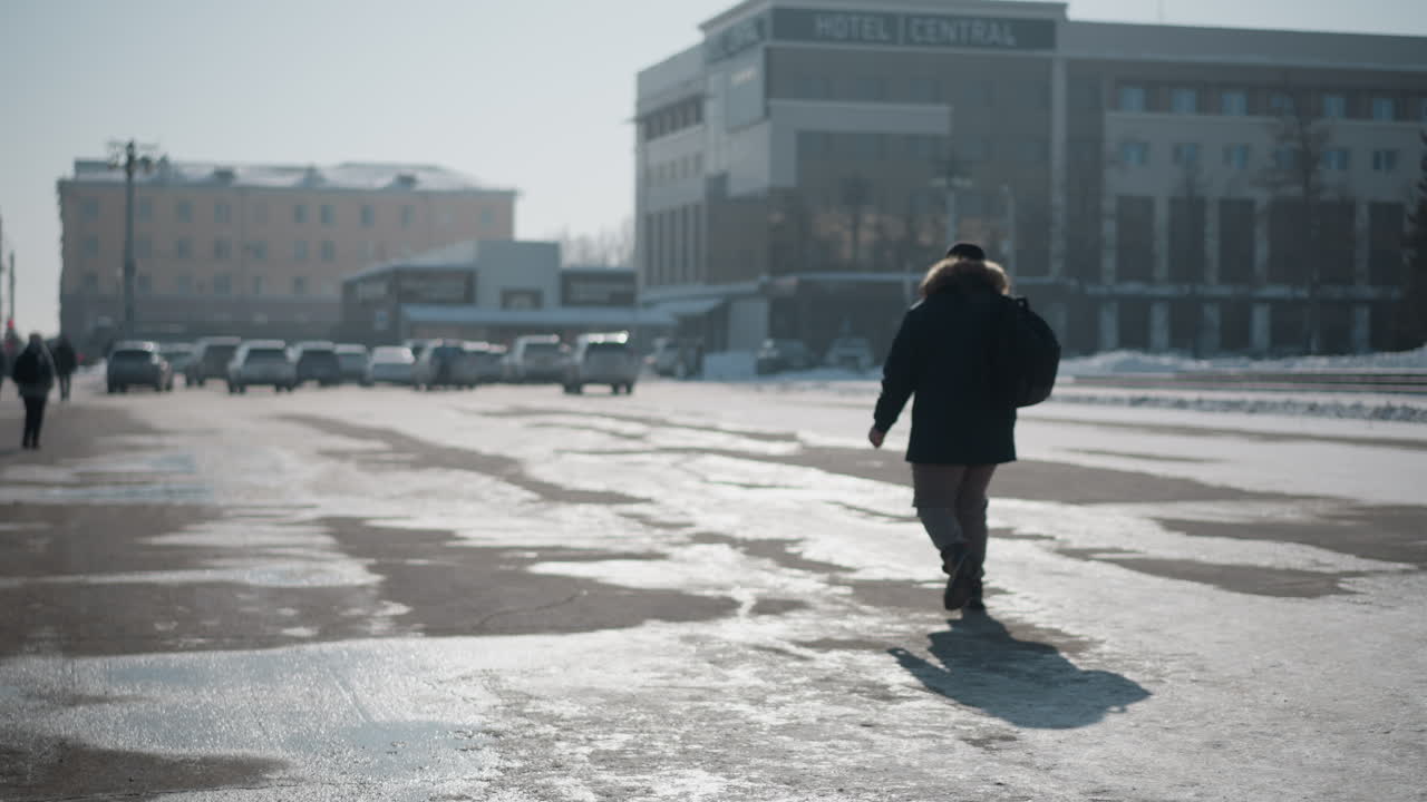 back view of young man with bag walks across icy parking toward hotel, long shadow trailing over wet pavement, winter sun glinting, cars and buildings ahead set calm urban morning commute mood