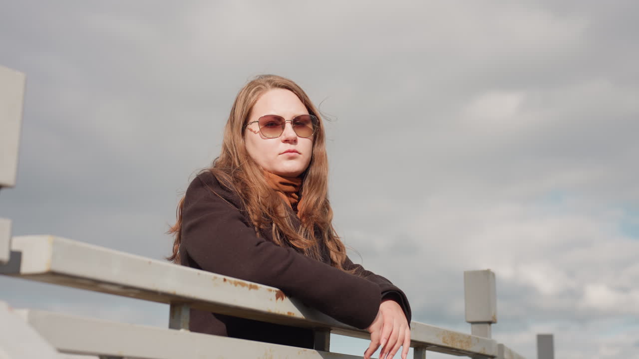 American tourist with long hair wearing dark suit and brown scarf leans on metal railing, glowing under daylight with gentle breeze, expressing calm freshness, outdoor relaxation, and peaceful mood