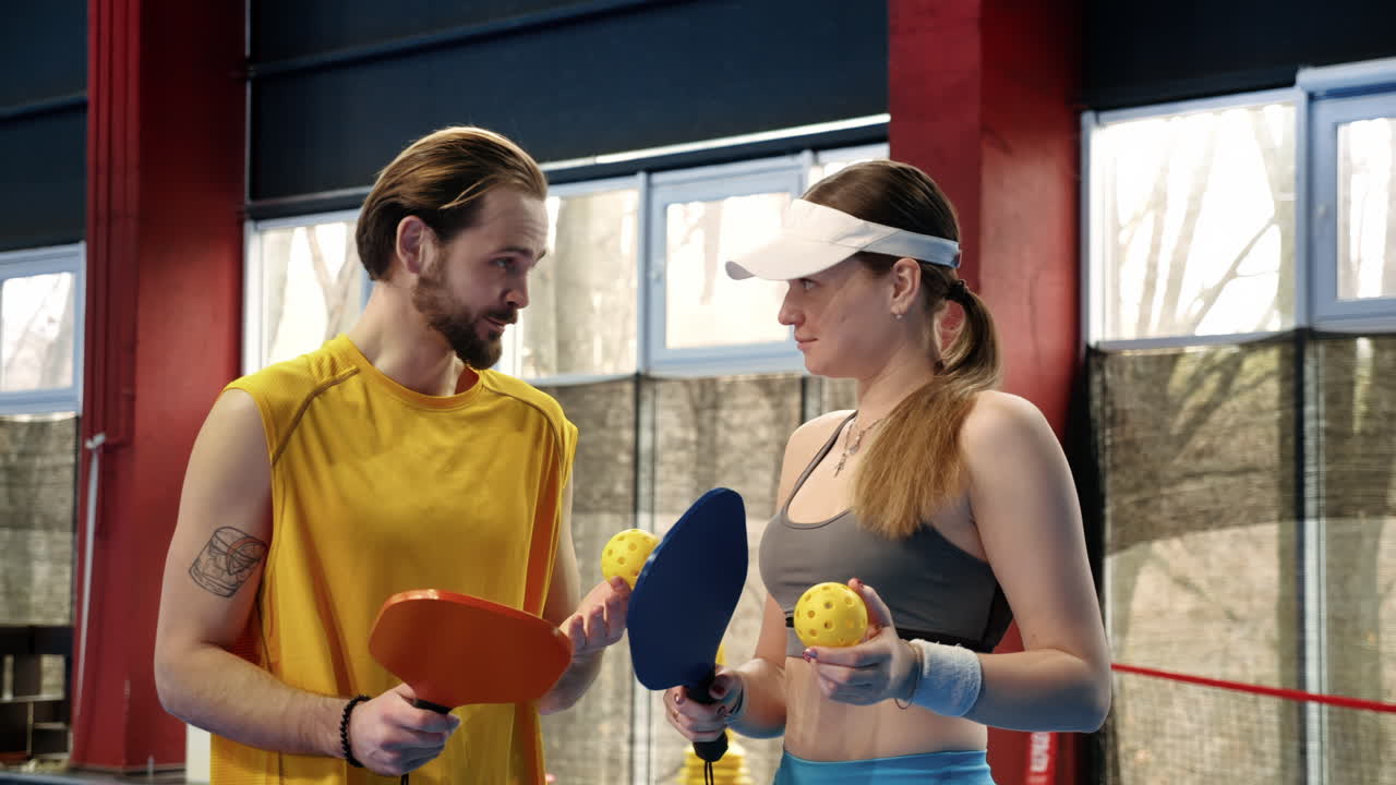 A man and a woman talking and looking at paddles after playing pickleball on a blue, inside court