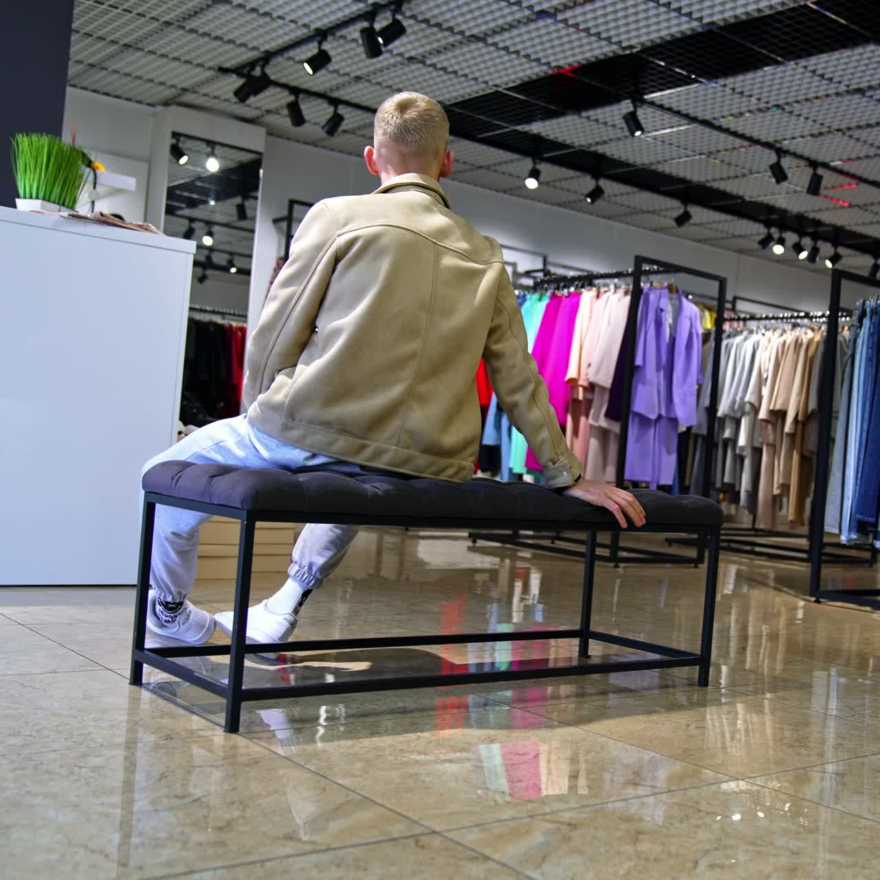 Blond man in leather jacket sits his back to the camera in the clothes shop. Person is waiting for somebody at the fitting room nervously tapping his fingers and foot