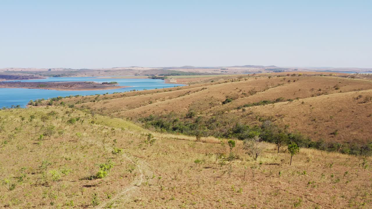 Drone orbits Grotas no Cerrado in Goiás revealing dry rolling hills, sparse vegetation, and a blue reservoir with islands showing biome zoning and terrain-water contrast in Brazil’s central savanna