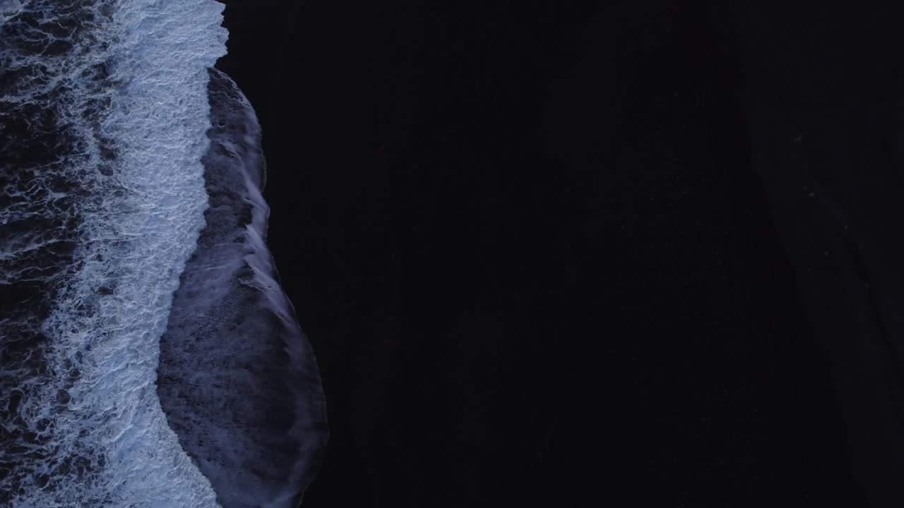 A striking vertical drone shot presents an abstract close-up of a vibrant wave meeting the distinctive black volcanic sand of Reynisfjara Beach in Iceland
